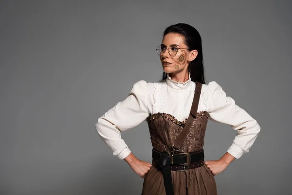 Nachdenkliche Steampunk-Frau mit Brille, die Hände auf den Hüften isoliert auf grau — Stockfoto