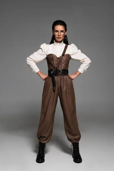 Full length view of steampunk woman in glasses standing with hands on hips on grey — Stock Photo