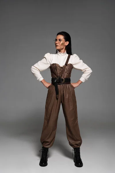 Vista completa de la mujer steampunk pensativa en gafas de pie con las manos en las caderas en gris - foto de stock