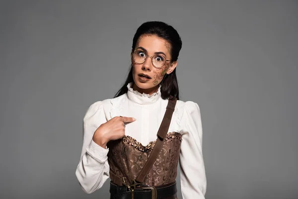 Vista frontal de la sorprendida mujer steampunk apuntando con el dedo a sí misma aislada en gris - foto de stock