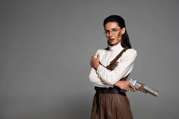 Donna steampunk pensierosa in bicchieri che tiene la pistola e distoglie lo sguardo sul grigio — Foto stock