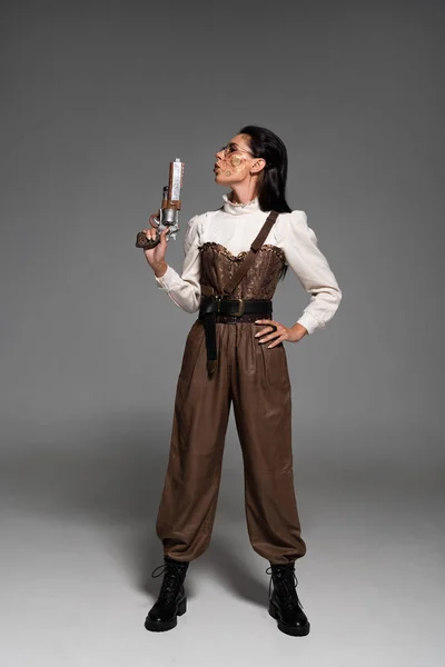 Visão comprimento total da mulher steampunk confiante de pé com a mão no quadril e segurando a pistola no cinza — Fotografia de Stock