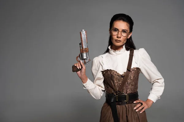 Attraente donna steampunk in piedi con mano sul fianco e tenendo pistola isolata sul grigio — Foto stock