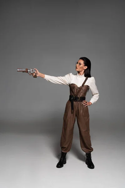 Vista completa de la atractiva mujer steampunk de pie con la mano en la cadera y apuntando con pistola en gris — Stock Photo