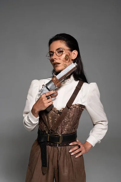 Attractive steampunk woman standing with hand on hip and holding pistol isolated on grey — Stock Photo