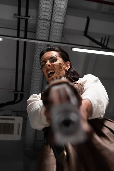 Selective focus of angry steampunk woman screaming and aiming with pistol at camera — Stock Photo
