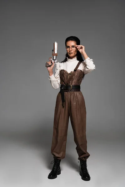 Full length view of steampunk woman holding vintage pistol and touching glasses on grey — Stock Photo