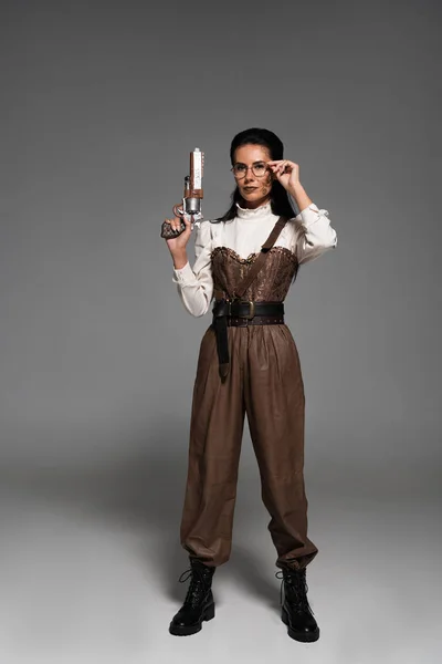 Visão de comprimento total da mulher steampunk segurando pistola vintage e óculos tocantes no cinza — Fotografia de Stock