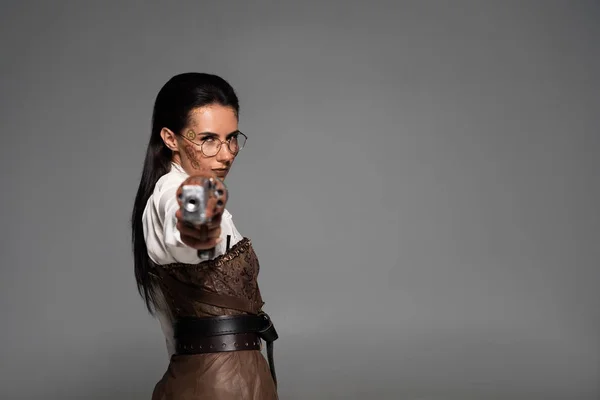 Selective focus of confident steampunk woman aiming with pistol at camera isolated on grey — Stock Photo