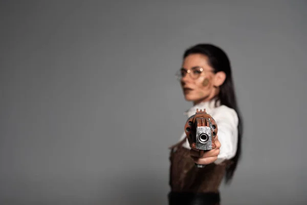 Селективный фокус уверенной стимпанк-женщины, нацеленной с пистолетом на камеру, изолированную на сером — стоковое фото