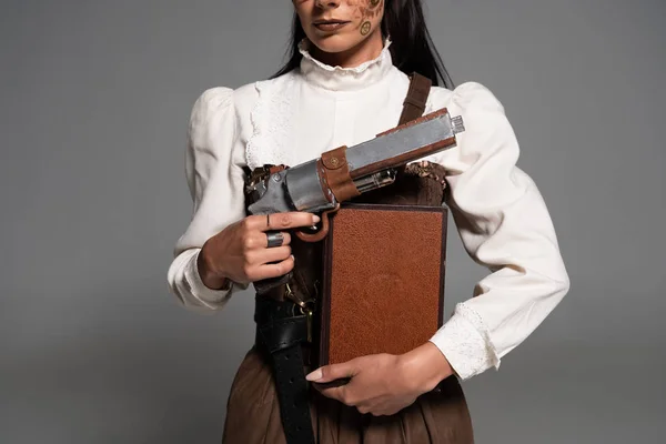 Cropped view of steampunk woman holding book and vintage pistol on grey — Stock Photo