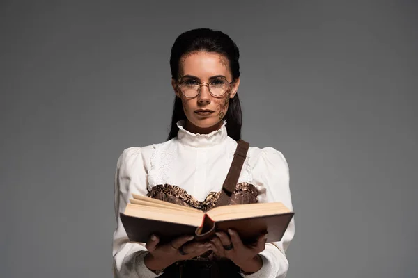 Frontansicht einer Steampunk-Frau mit Brille, die ein aufgeschlagenes Buch isoliert auf grau hält — Stockfoto