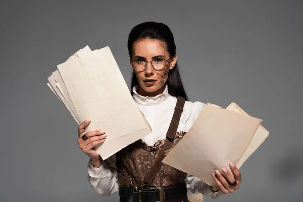 Attractive steampunk woman in glasses holding documents and looking at camera on grey — Stock Photo