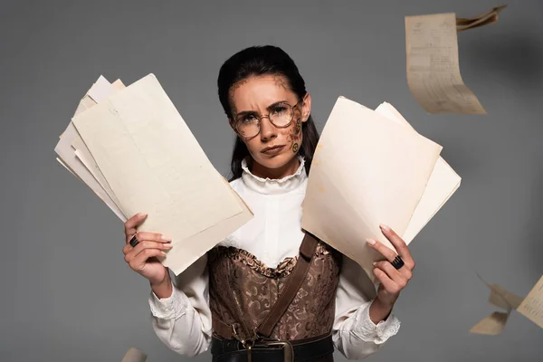 Displeased steampunk woman in glasses holding documents and looking at camera on grey — Stock Photo