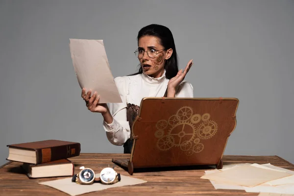 Inquiet steampunk femme assise à table avec ordinateur portable vintage et des livres et en regardant des documents isolés sur gris — Photo de stock
