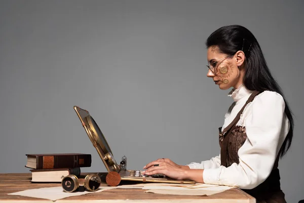 Vista laterale di focalizzata donna steampunk attraente utilizzando laptop sul posto di lavoro isolato sul grigio — Foto stock