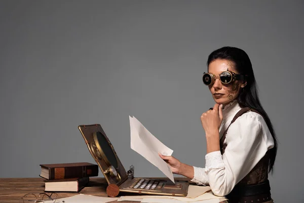 Стимпанк женщина с документами за столом с винтажным ноутбуком изолированы на серый — стоковое фото