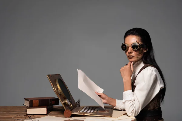 Steampunk woman holding documents at table with vintage laptop isolated on grey — Stock Photo