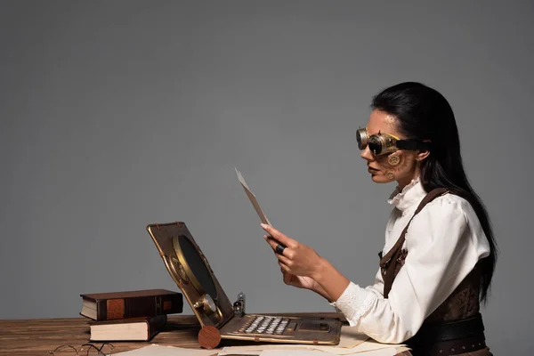 Steampunk woman holding document at table with vintage laptop isolated on grey — Stock Photo