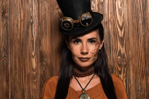 Vue de face de la femme steampunk en chapeau haut de forme avec lunettes regardant la caméra sur bois — Photo de stock