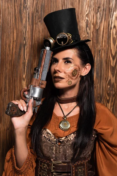 Mulher steampunk pensativo no chapéu superior com óculos segurando pistola em madeira — Fotografia de Stock