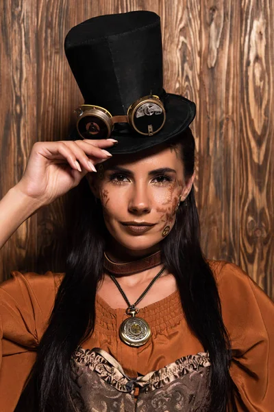 Front view of steampunk woman touching top hat with goggles looking at camera on wooden — Stock Photo