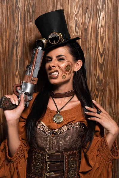 Irritated steampunk woman in top hat with goggles holding pistol on wooden — Stock Photo