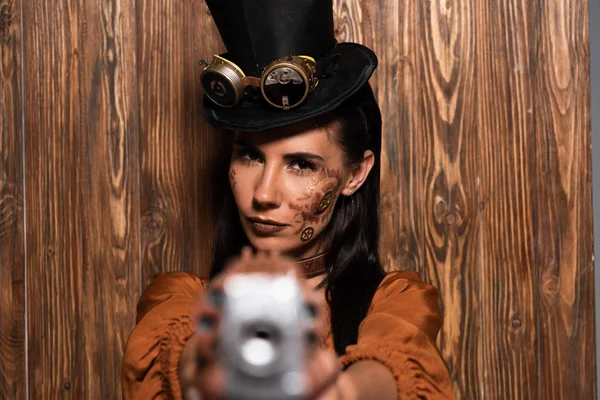 Foyer sélectif de la femme steampunk grave dans le chapeau supérieur avec des lunettes visant avec pistolet à la caméra sur bois — Photo de stock