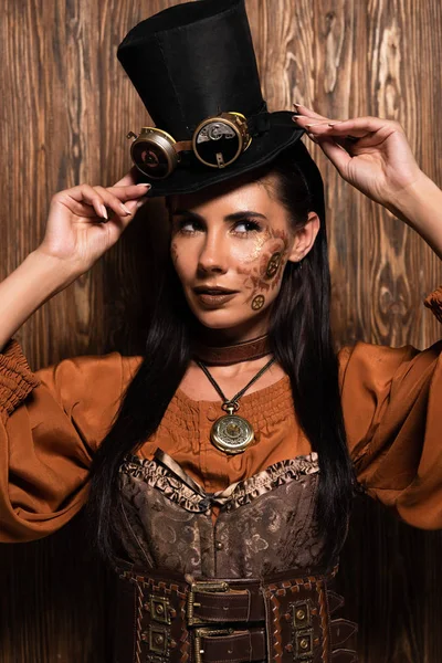 Attractive steampunk woman touching top hat with goggles on wooden — Stock Photo