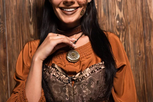 Vista recortada de mujer sonriente con maquillaje steampunk tocando medallón en madera - foto de stock