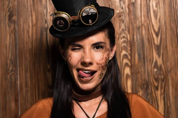 Front view of smiling steampunk woman blinking and sticking out tongue on wooden — Stock Photo