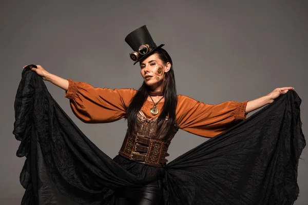 Atractiva mujer steampunk en sombrero de copa con gafas jugando con falda aislada en gris - foto de stock