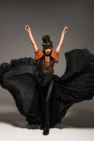 Vue pleine longueur de femme steampunk attrayant dans le chapeau supérieur avec des lunettes debout avec les mains vers le haut sur gris — Photo de stock