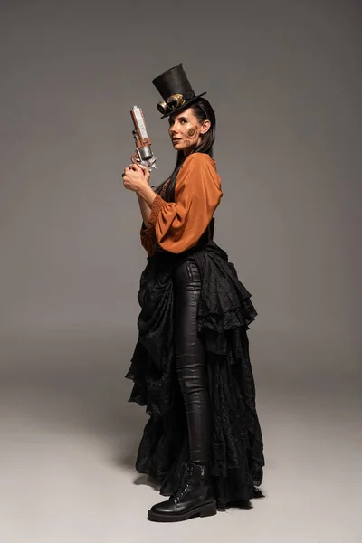 Вид в полный рост женщины-стемпанка в верхней шляпе с когтями, держащими пистолет на сером — стоковое фото