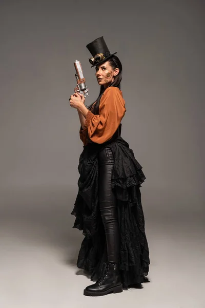 Full length view of steampunk woman in top hat with goggles holding pistol on grey — Stock Photo