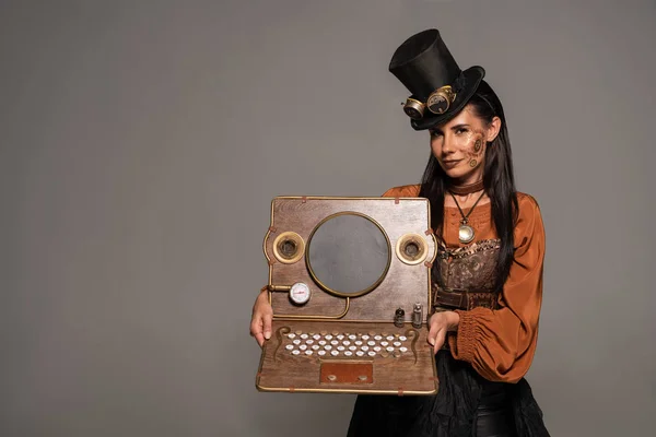 Smiling woman in top hat with goggles showing steampunk laptop isolated on grey — Stock Photo