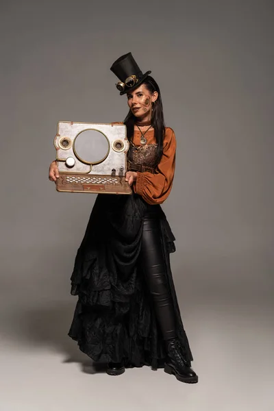 Full length view of woman in top hat with goggles showing steampunk laptop on grey — Stock Photo