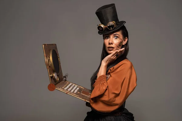 Шокована жінка у верхній шапці з окулярами, використовуючи стімпанк ноутбук ізольовано на сірому — стокове фото