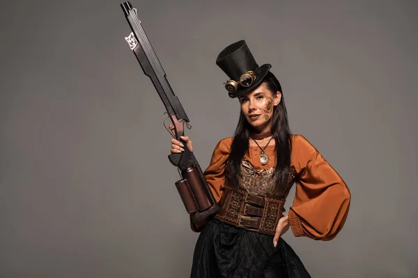 Привлекательная стимпанк-женщина в цилиндре с пистолетом в руке на бедре, изолированная на сером — стоковое фото