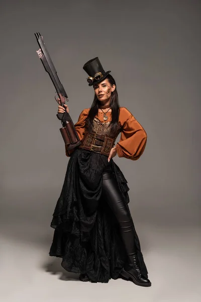 Vista completa de la atractiva mujer steampunk en top hat sosteniendo pistola y de pie con la mano en la cadera en gris - foto de stock