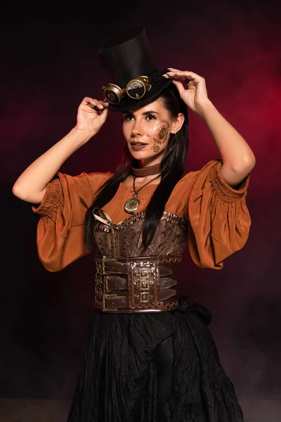 Attractive steampunk woman with makeup putting on top hat on black in pink smoke — Stock Photo