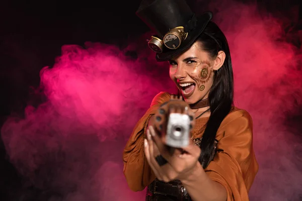 Screaming steampunk woman aiming with pistol to camera in pink smoke on black — Stock Photo