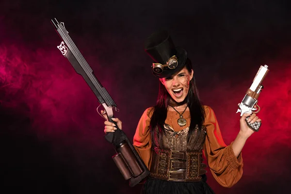 Vista frontal de la mujer steampunk gritando sosteniendo arma y pistola en humo rosa en negro - foto de stock