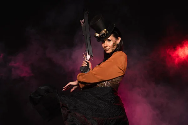 Attractive steampunk woman in top hat holding gun in pink smoke on black — Stock Photo