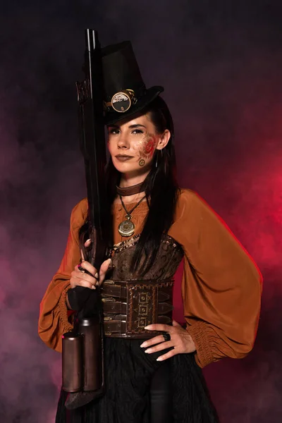 Attractive steampunk woman holding gun and standing with hand on hip in pink smoke — Stock Photo