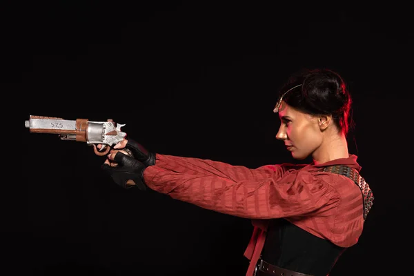 Side view of confident steampunk young woman holding pistol isolated on black — Stock Photo