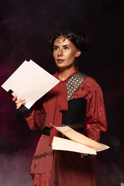 Pensive attractive steampunk young woman holding papers in smoke on black — Stock Photo