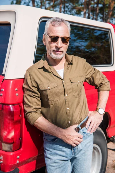 Confident senior man in sunglasses standing with hand on hip near red car and looking at camera — Stock Photo