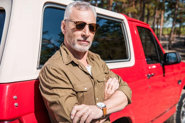 Homme âgé confiant dans des lunettes de soleil debout avec les bras croisés près de voiture rouge et regardant la caméra — Photo de stock