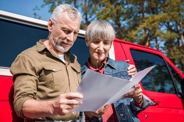 Pareja de viajeros mayores de pie cerca de coche rojo, abrazando y sosteniendo el mapa en un día soleado - foto de stock